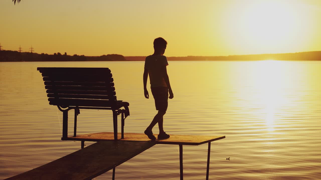 Silhouette of a boy goes on the bridge over the lake and sees at the sunset. Slow motion of the kid on empty bridge walking to watch a beautiful sunset near the river