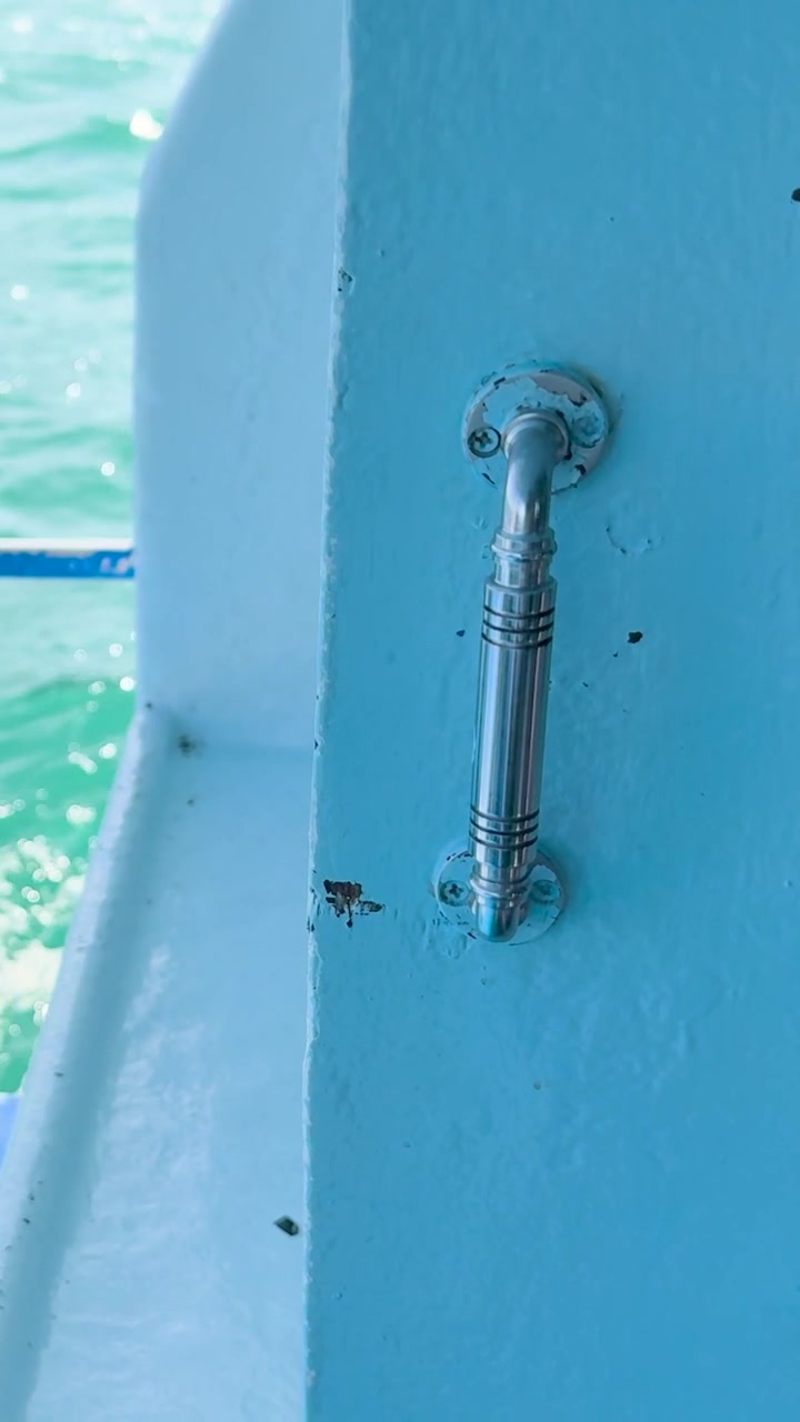 Uno scatto verticale che si concentra su una maniglia cromata, con una barca blu e il mare turchese sullo sfondo.