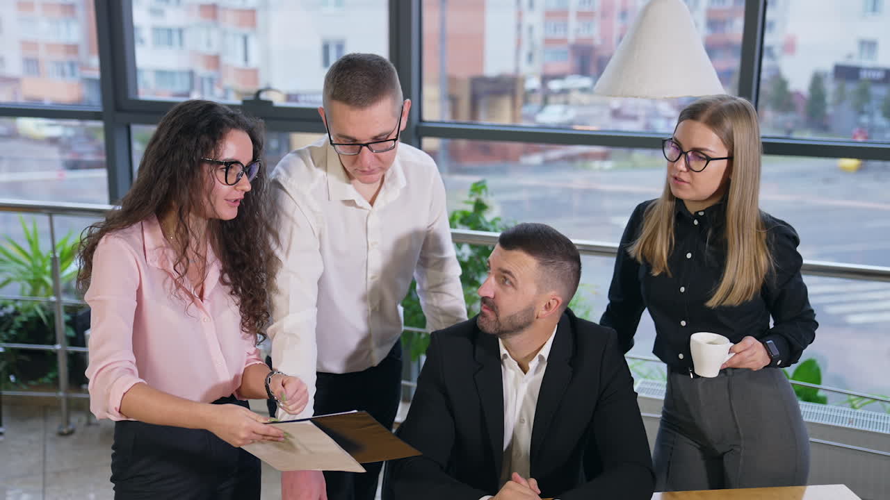 Female with long dark curly hair talks to colleagues and shows the paper. Office people came to hear the new ideas from a teammate.