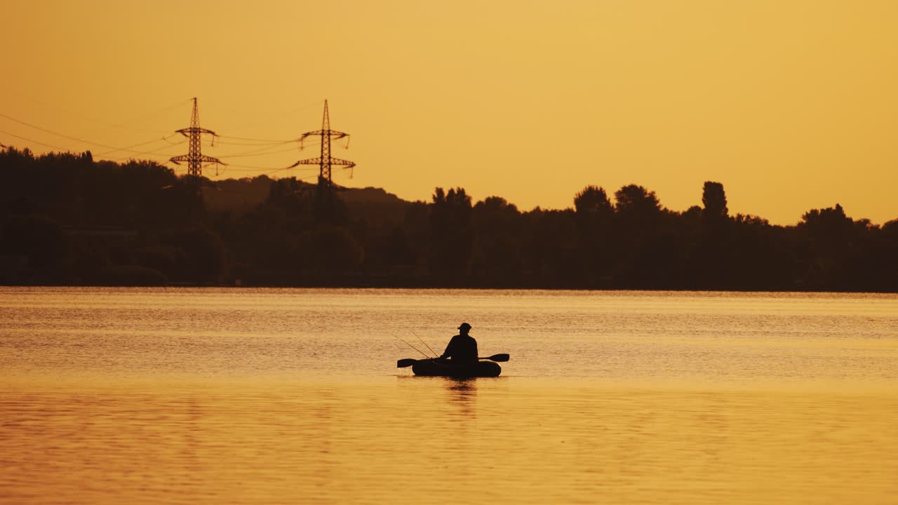 Silhouette of a man floating in a boat on the river surface on the background of an evening trees. Fisherman roams with oars while sailing on a row boat in the lake at beautiful sunset.