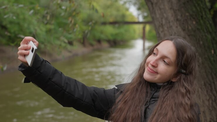 Avventuriera che fotografa paesaggi fluviali. Viaggiatrice solitaria che scatta foto panoramiche vicino all'acqua. Giovane donna con capelli castani che esplora la natura e immortala splendidi paesaggi sulle rive del fiume.