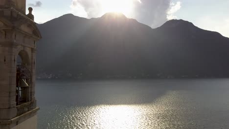 Epic-Church-Clock-Tower-Closeup-with-sun-rays-on-a-lake-with-mountains-in-the-background,-Nesso,-Lago-di-Como,-Italy