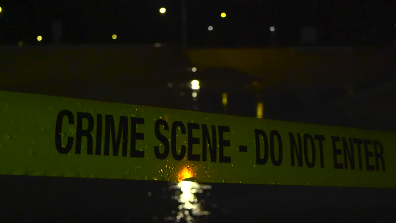 Cinta policial amarilla de escena del crimen con el texto 'No Pasar' frente a un skatepark nocturno durante una tarde lluviosa con gotas de lluvia cayendo sobre la cinta a cámara lenta. También se reflejan las luces de la calle.