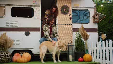 feliz chico moreno en una camisa a cuadros verde acaricia a su gran perro de color crema y una chica morena toca su cabello cerca de su remolque durante un picnic en un campamento fuera de la ciudad en el verano