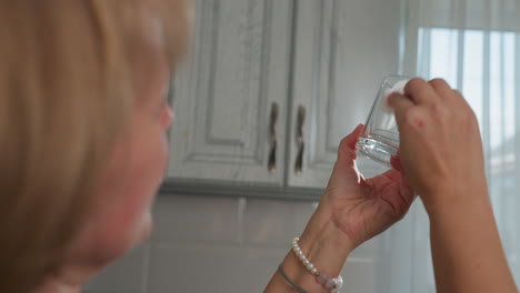 close up of lady using tissue paper to carefully dry glass cup while inspecting its cleanliness near kitchen cabinet with natural light shining through window to ensure sparkling spotless surface