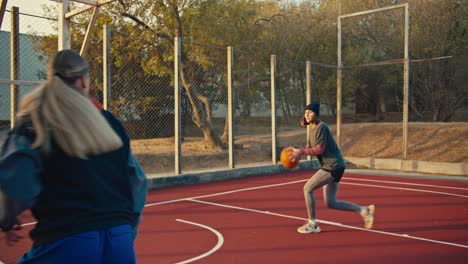 A-blonde-girl-in-a-sports-uniform-gives-a-pass-with-an-orange-sword-to-her-friend-who-throws-the-ball-into-the-basket-during-their-game-of-basketball-on-a-red-street-court-in-the-morning