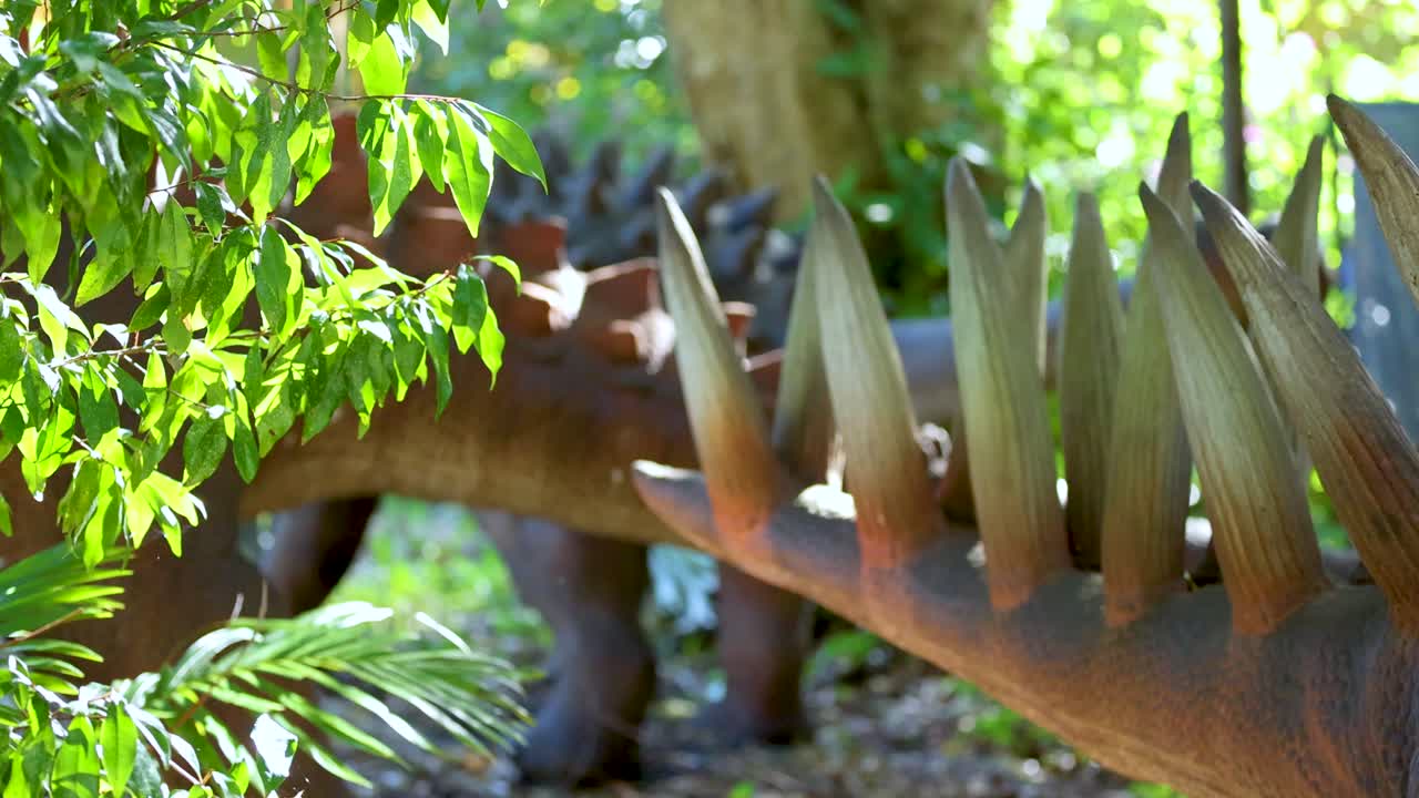 A Kentrosaurus model stands amidst vibrant greenery in a dinosaur park, captured with dynamic camera movement and natural lighting
