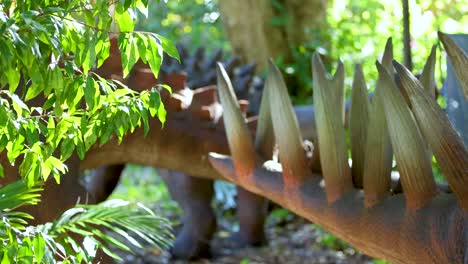 a kentrosaurus model stands amidst vibrant greenery in a dinosaur park, captured with dynamic camera movement and natural lighting