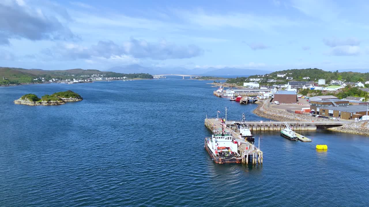 Aerial footage moves steadily over blue water toward a pier with docked boats, a coastal town, and distant green hills under daylight