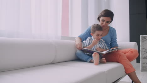 Madre-Amorosa-Enseñando-A-Su-Hijo-A-Leer-Sosteniendo-Un-Libro-Hablando-En-El-Sofá-En-Casa