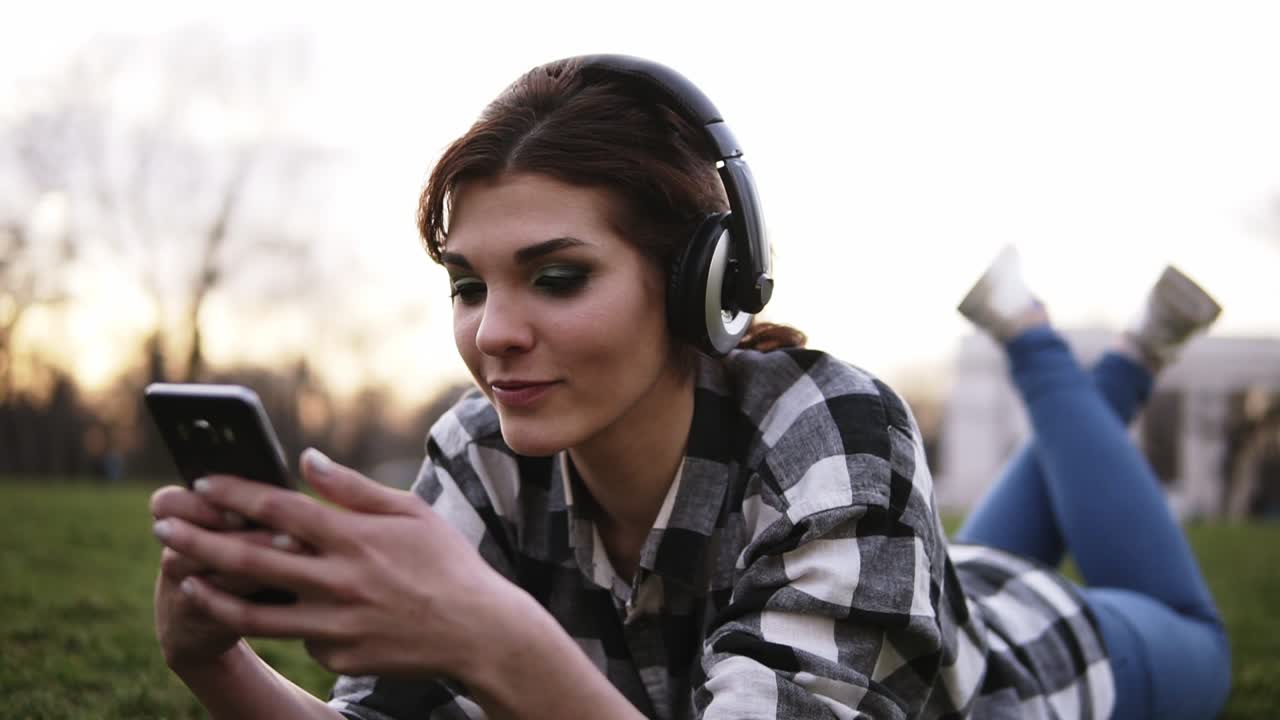 chica joven con auriculares elegantes acostada en el césped, escuchando música y hablando en el móvil.