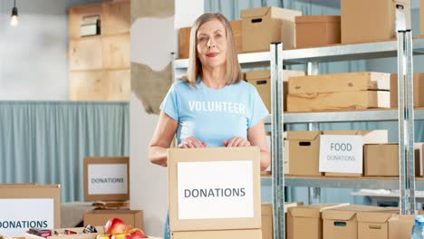 Mujer-Caucásica-Voluntaria-Empacando-Cajas-De-Donación-Con-Manzanas-Rojas-Y-Mirando-La-Cámara-En-Un-Almacén-De-Caridad