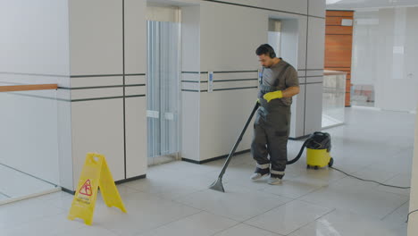 Arabic-Cleaning-Man-Wearing-Headphones-Cleaning-With-Pressure-Water-Machine-While-Dancing-Inside-An-Office-Building-Behind-A-Wet-Floor-Warning-Sign