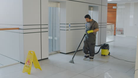 hombre de limpieza árabe con auriculares limpiando con máquina de agua a presión mientras baila dentro de un edificio de oficinas detrás de una señal de advertencia de suelo mojado