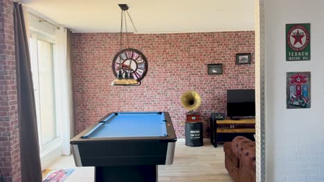 a bright, vintage-inspired apartment living space with a billiard table, exposed brick wall, large clock, and eclectic antique decor. smooth daylight pan shot