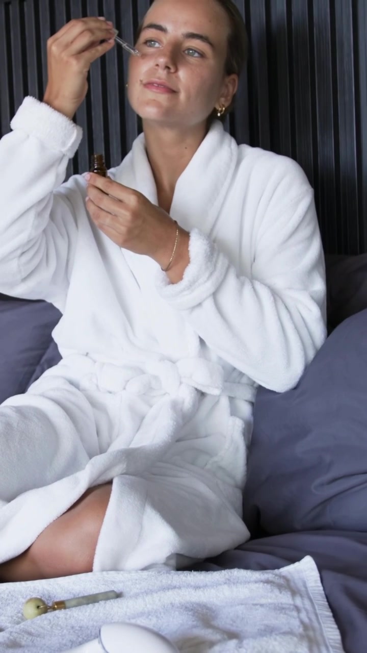 Vertical video: Drawing from amber dropper, woman in bathrobe applying serum in bed to hydrate skin