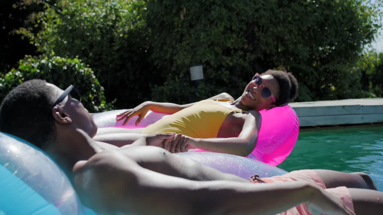 African American couple holding hands, relaxing on backyard pool floats, woman in yellow swimsuit