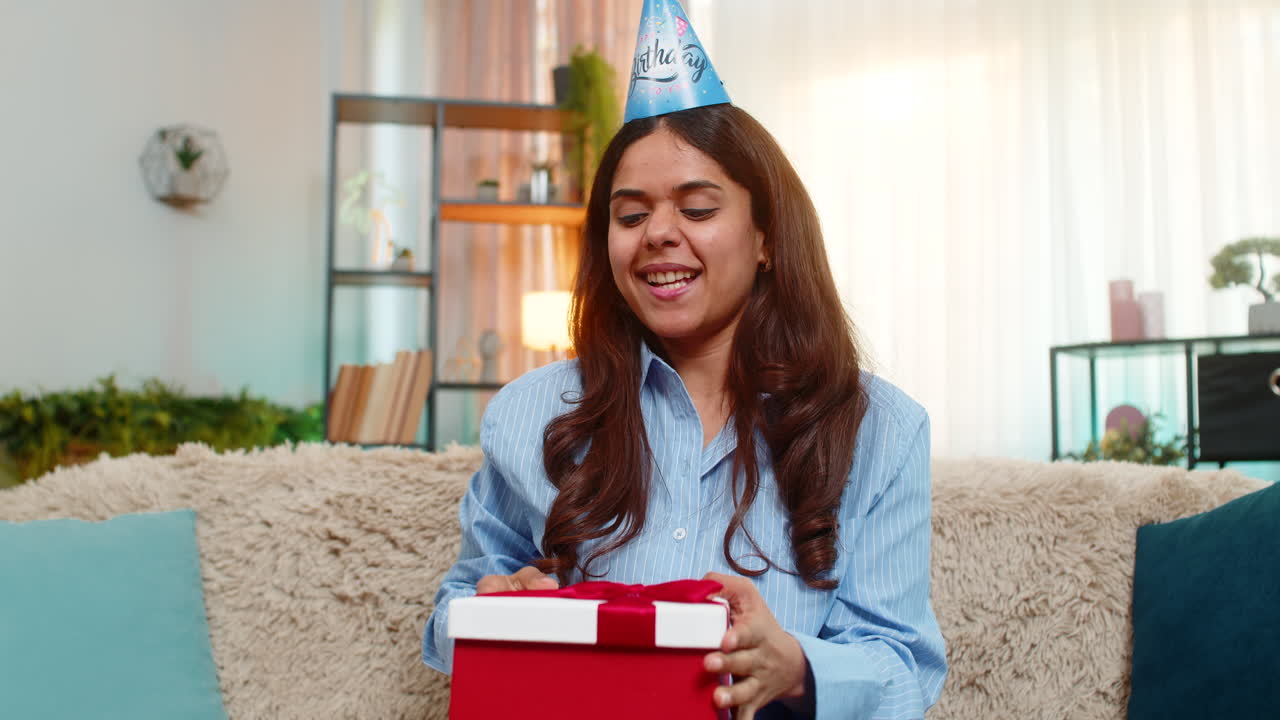 Mujer india abriendo un regalo de cumpleaños en el sofá de casa reaccionando con sorpresa y alegría sonriendo de alegría