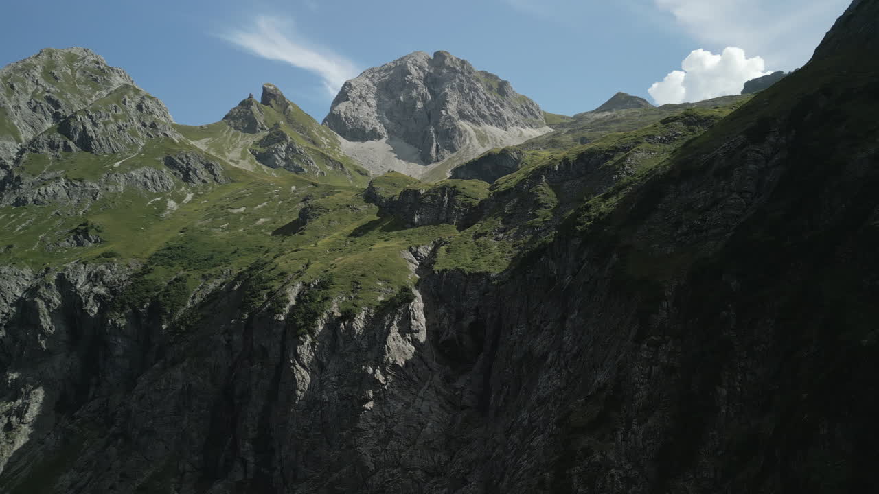 Drone aerial view on the edge of a rocky mountain looking upwards towards the blue sky with clouds. The camera turns towards the summit.