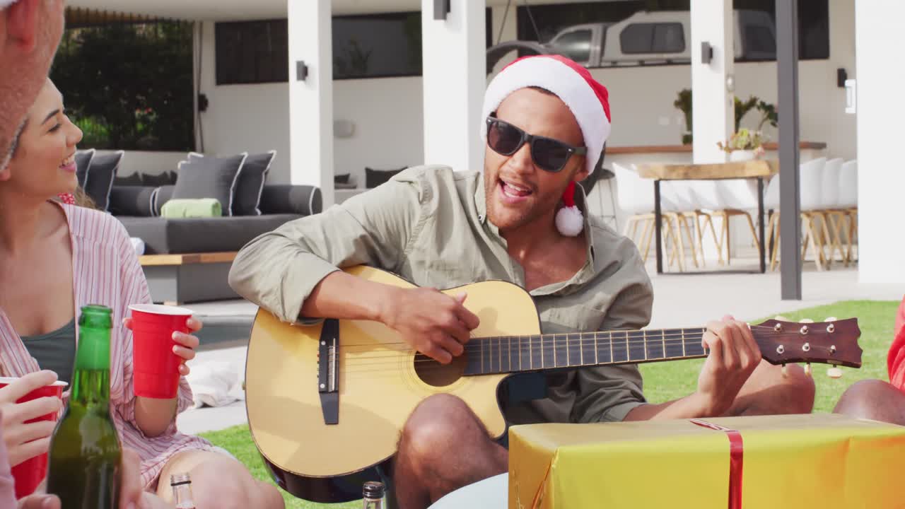 Gruppo felice di amici di diverse etnie che suonano la chitarra a una festa di Natale in giardino