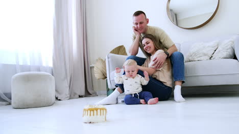 loving parents sit watching their adorable baby. little kid is busy with some spot on the floor not paying attention to the cake in front of him.