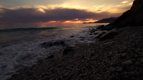 dark clouds on red sunset sky reflecting over sea surface seen from the coast washed by waves in mediterranean