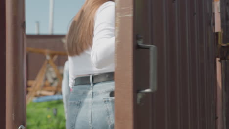 Rear view of lady wearing denim jacket opening brown metal gate as friends follow behind, sunny outdoor setting with soft shadows, emphasizing motion, action, and casual group interaction