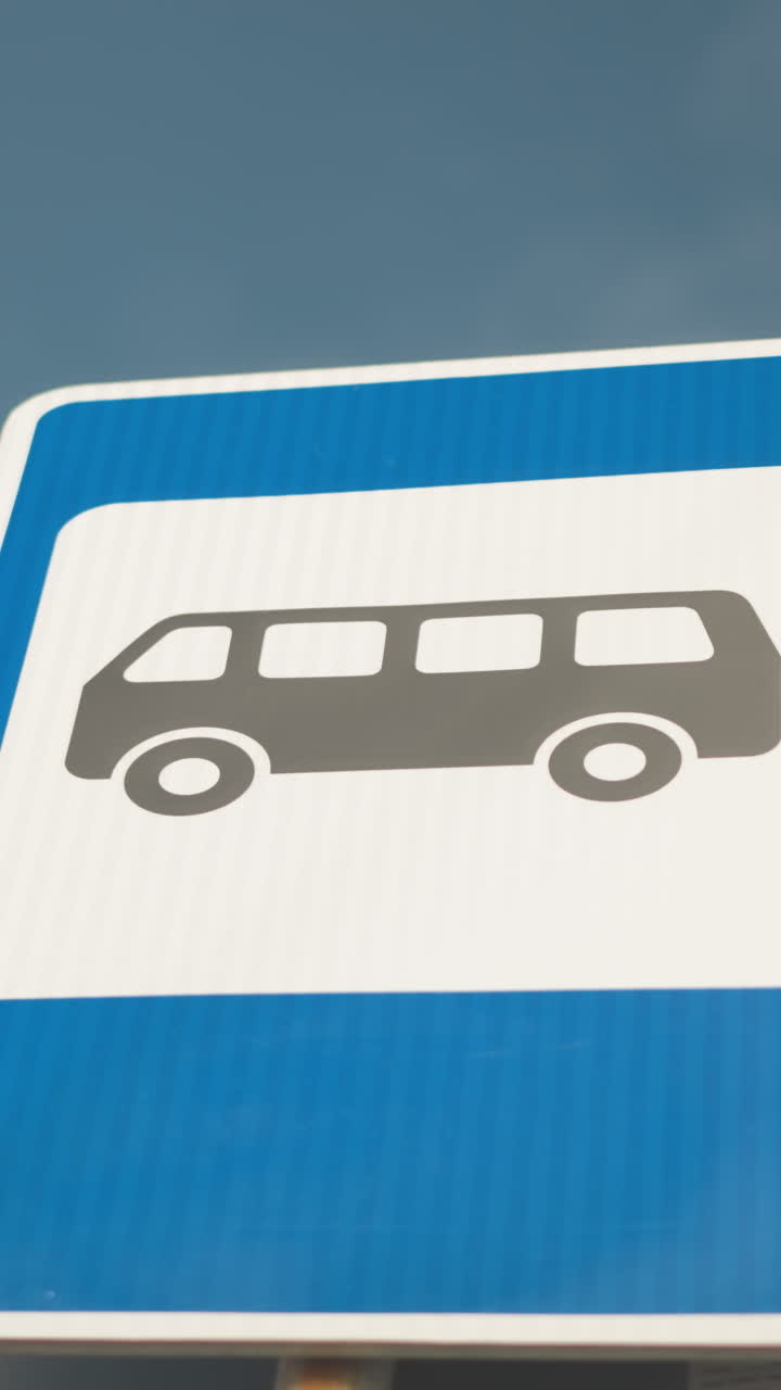 Primer plano de una señal de autobús en ángulo contra el cielo; panel azul vibrante con un borde blanco nítido y silueta de autobús gris; perspectiva inclinada definida que captura el poste de metal reflectante; estética moderna y limpia; ideal