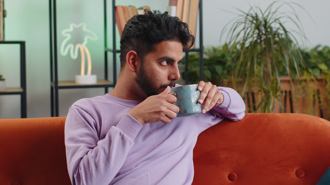 웃는 인도인 남자가 오렌지 소파에서 아침에 따뜻한 커피 또는 허브 차 한 잔을 마시고 있습니다.