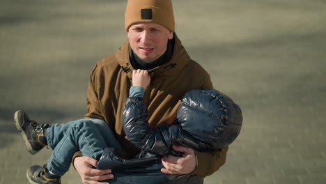 a close-up of father carrying his small child who is sleeping while walking, the father appears stressed, with the child's hand gently grasping his collar