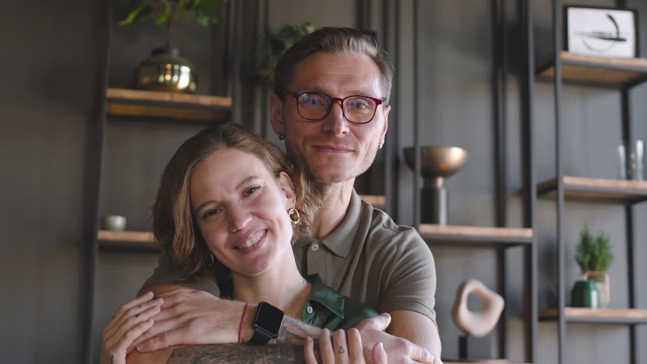 Portrait Of A Happy Couple Smiling At Camera At Home While Man Embracing Woman From Behind