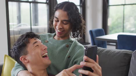 Happy-diverse-male-couple-sitting-on-sofa,-embracing-and-using-smartphone-in-living-room