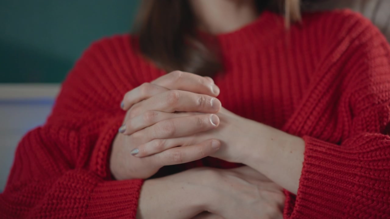 Hands remain clasped in supportive embrace while subtle shifting motion suggests ongoing comfort and reassurance, with red knit sleeves, soft indoor light and caring human connection
