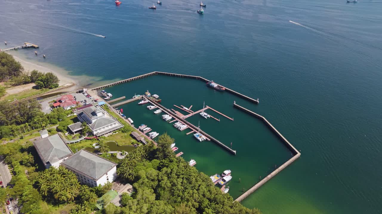 Slow push in drone shot of Victoria Jetty in Labuan, Malaysia, showing boats moored along turquoise waters beside lush greenery. Calm, tropical, and scenic harbor atmosphere