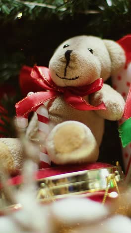 soft teddy bear toy with red ribbon rotating among christmas tree decorations in vertical composition