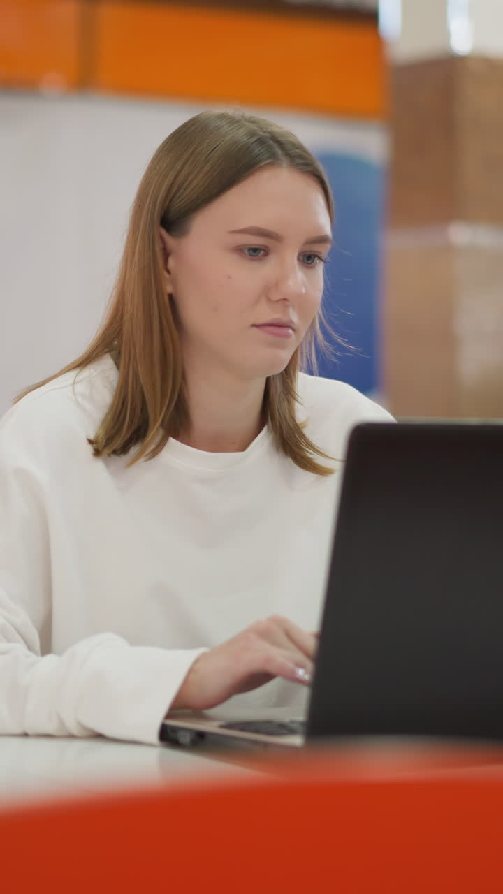 mujer trabajando remotamente en una computadora portátil en un restaurante, mirando momentáneamente hacia otro lado antes de reanudar el trabajo, decoración vibrante y fondo borroso con personas a su alrededor