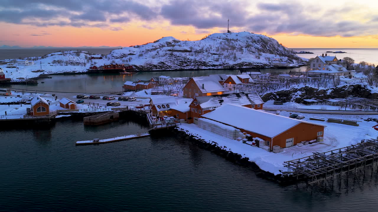 鮮やかな冬の夕焼けに染まるロフォーテン諸島の雪に覆われたノルウェーの漁村を捉えたシネマティックな空撮ドローン映像。北極海のほとりに建つ伝統的な黄色と赤の小屋が特徴。