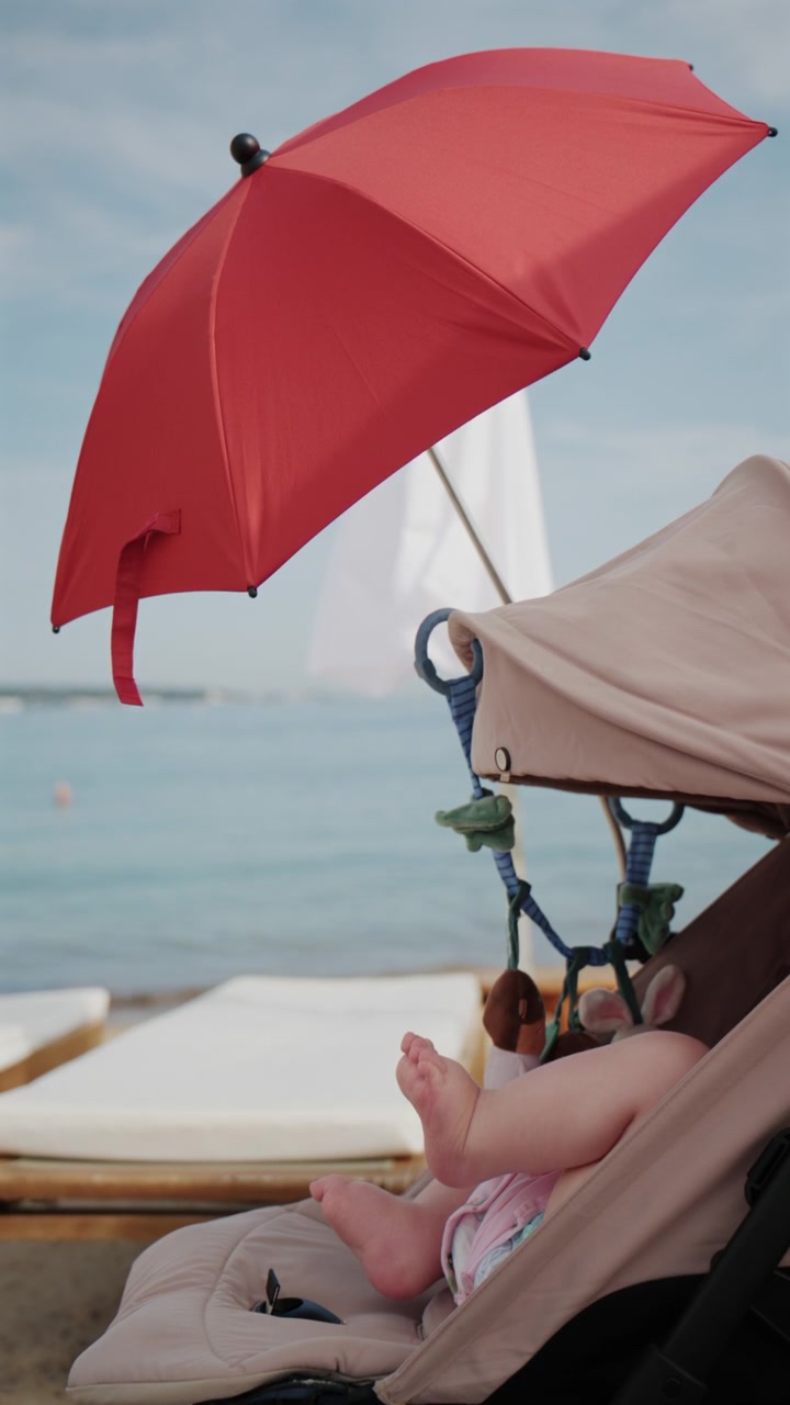 Bambino seduto nel passeggino sotto un ombrellone da spiaggia rosso brillante in riva al mare.