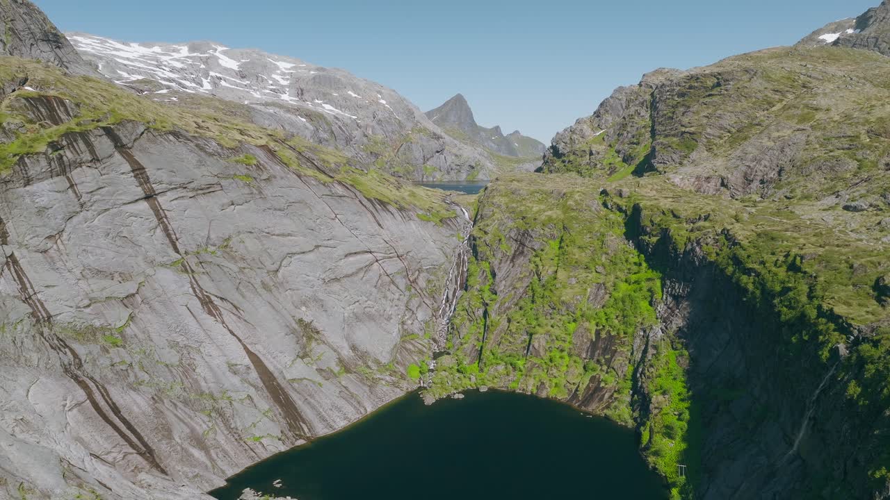 Un sereno lago blu intenso si trova sotto ripide scogliere di granito, mentre una cascata scende da un lago alpino più alto, circondato da cime innevate nelle Lofoten, Norvegia.