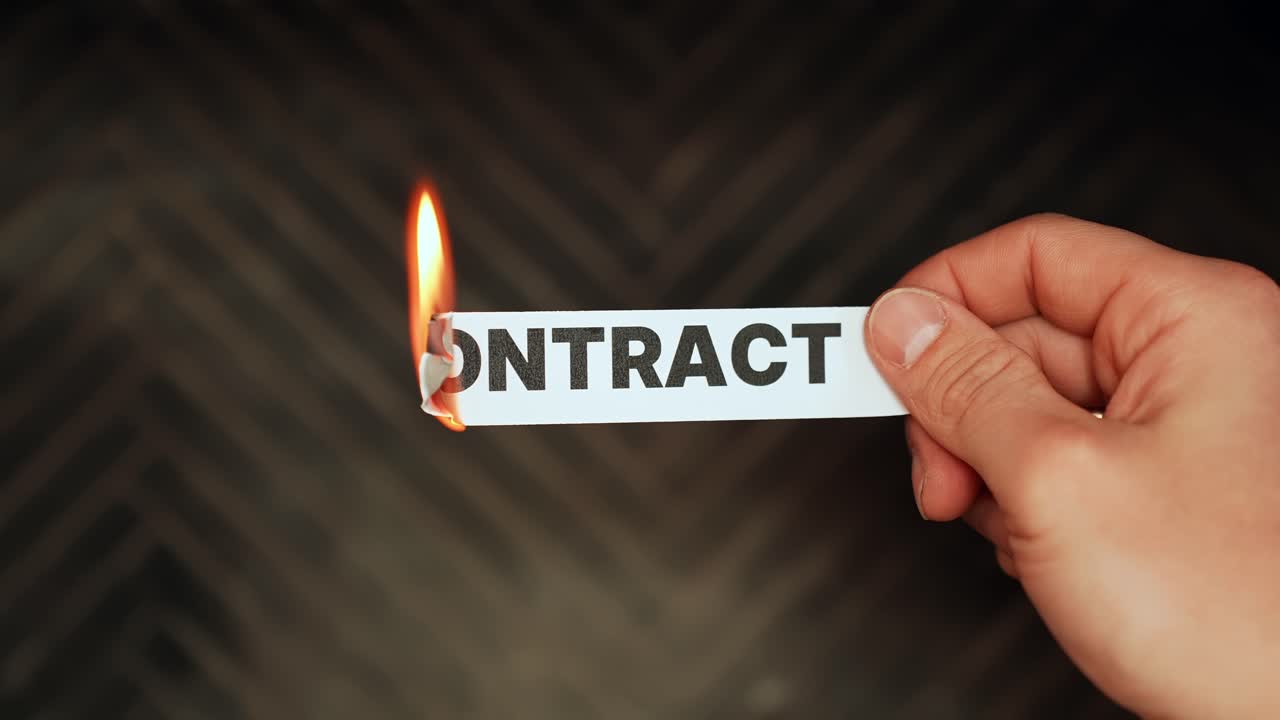 Close-up of a hand holding a paper strip labeled “CONTRACT” while the edge burns, symbolizing agreement risk, breach, termination, and legal fallout on a dark background with copy space