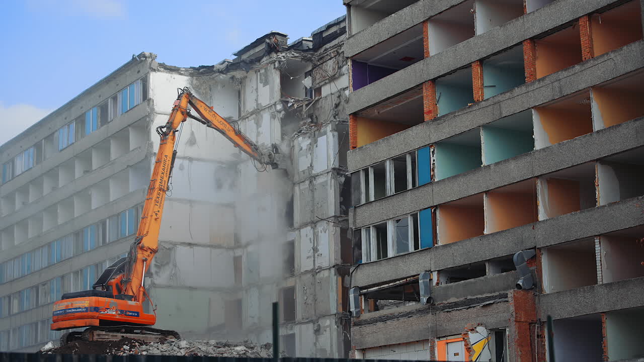 Compact orange demolishing machine dismantling the house. Deconstruction of a building ruined by the nature disaster. View from below.
