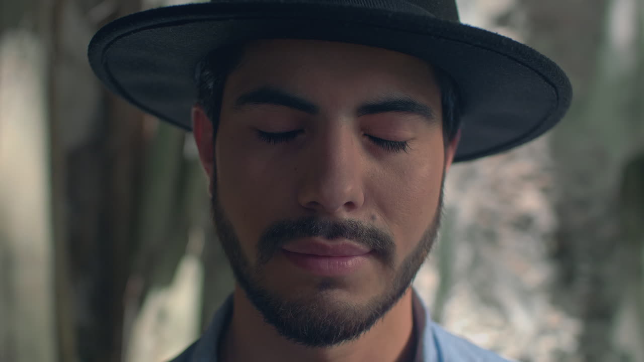 High-quality 4K footage of a stylish young man with a beard and hat, slowly opening his eyes in soft, natural light. He has a focused, mysterious expression. Ideal for travel, discovery