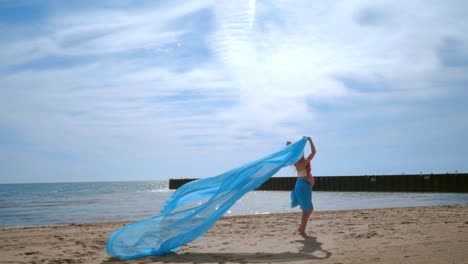 woman with pregnant belly holding blue fabric flying on wind at beach