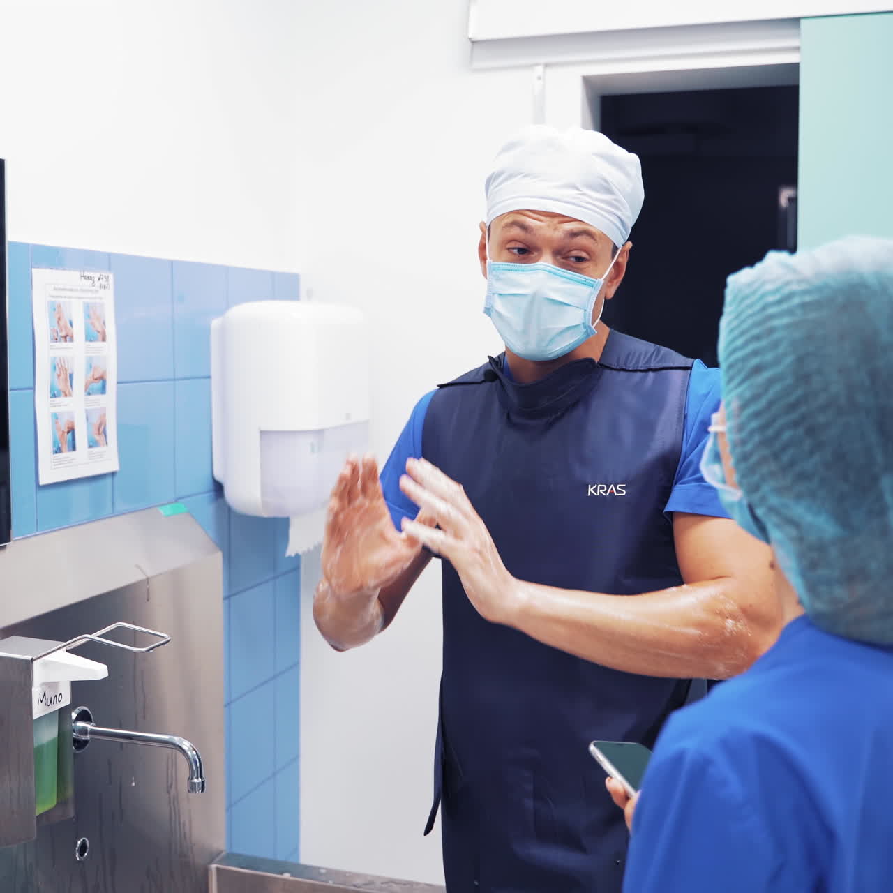 Doctor is chatting to a woman and washing his hands after the operation. Two doctors are talking in the bathroom in clinic. Surgeon makes his hands clean with a soap and water.