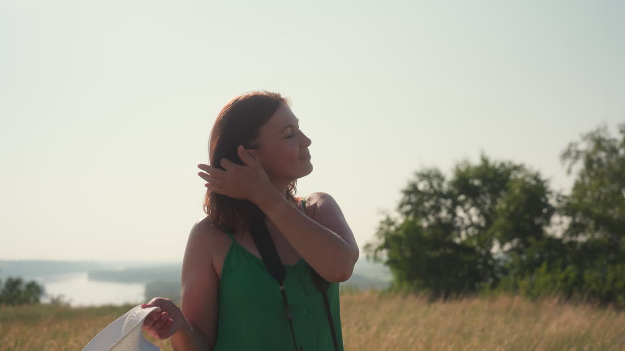 woman in green dress lifts wide hat from head while standing in sunlit dry grassy field, camera strapped on neck, gently adjusting hair with fingers, enjoying serene nature view on bright warm day