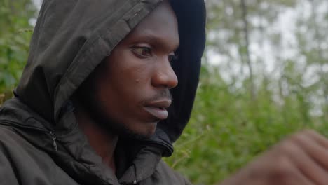 a close up shot of an african man wearing a hooded coat sitting down in a forest in africa