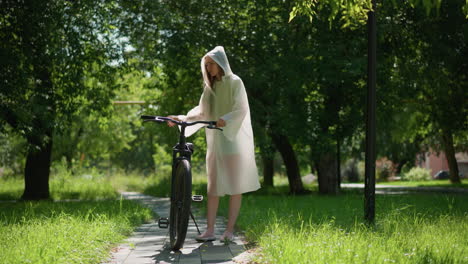 young lady in translucent raincoat strolls along sunny pathway with bicycle, stopping to place stand near pole, lowers hood, and leans on handlebars, surrounded by lush greenery and trees