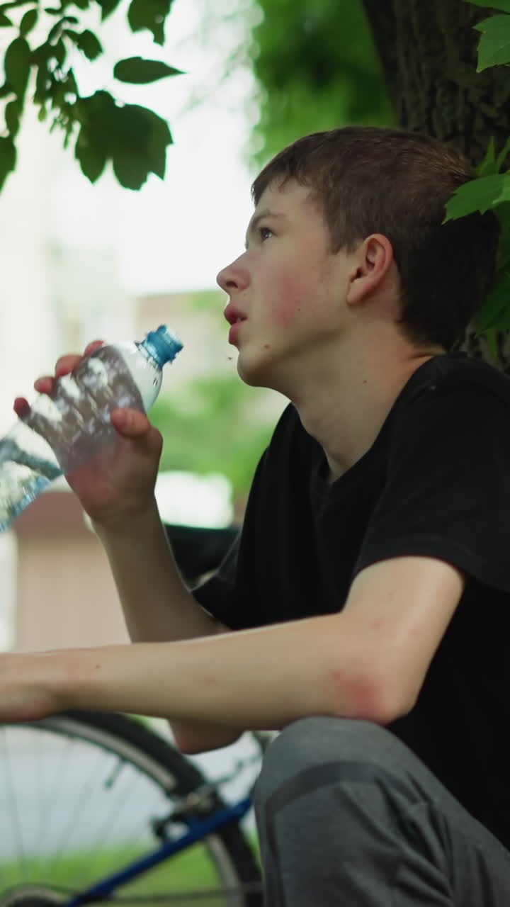 어린 소년은 주차된 자전거 의 나무 에서 휴식을 취하면서 물을 마시며, 거리를 가로지르는 건물의 흐릿한 시야와 함께 차갑게 목을 만집니다.