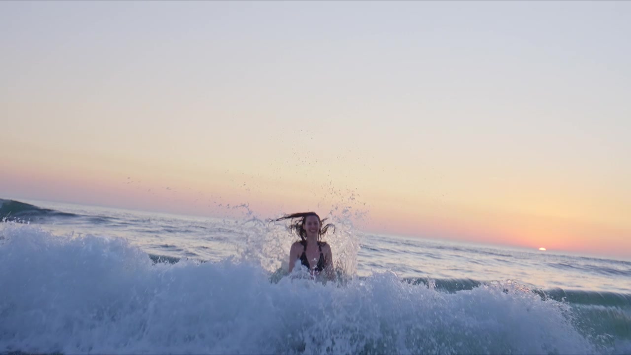 Giovane donna che cammina tra le onde al tramonto mentre un'onda che si infrange le si rovescia intorno, creando un'immagine drammatica e giocosa di stile di vita da spiaggia estivo