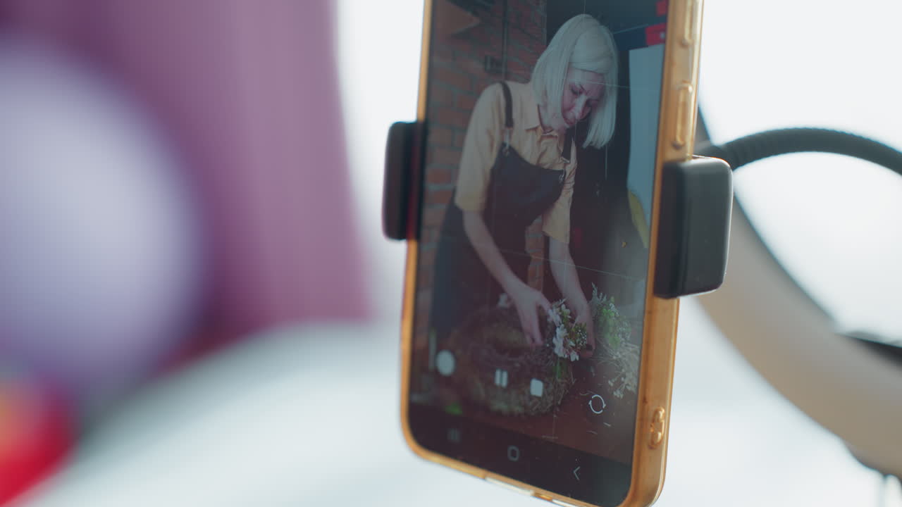 Smartfon z bliska na statywie z lampą pierścieniową nagrywa florystkę układającą wieniec, pokazując proces twórczy, transmisję na żywo, tworzenie zasobów, produkcję wideo do mediów społecznościowych w jasnym studio